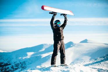 A person wearing a black ski suit, helmet and goggles on top of a mountain holding a red snowboard above their head. - CUF55362