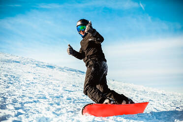 A person wearing a black ski suit, helmet and goggles skiing down a mountain on a red snowboard. - CUF55363