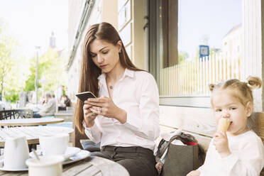 Mutter und Tochter sitzen vor einem Café, die Mutter telefoniert mit ihrem Handy, die Tochter isst ein Eis. - CUF55372