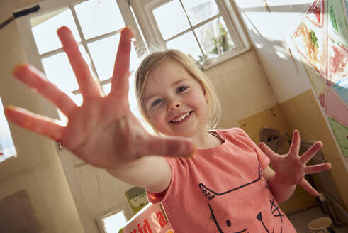 Smiling girl indoors during Corona virus crisis, doing finger painting, looking at camera. - CUF55388