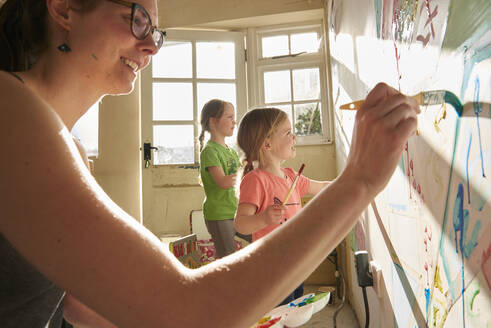 Lächelnde Frau und zwei Mädchen in einem Haus während der Corona-Viruskrise, Gemälde an einer Wand. - CUF55389