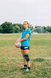 Eine Frau in Shorts und T-Shirt mit Knieschlaufe steht auf einem Trainingsplatz und hält einen Rugbyball. - CUF55394