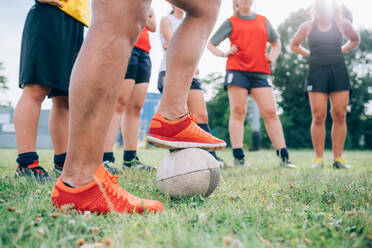 Die Beine und Füße einer Gruppe von Frauen, die auf einem Trainingsplatz stehen, eine mit einem Fuß auf einem Rugbyball. - CUF55399