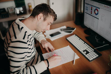 Ein Mann, der während des Einschlusses zu Hause arbeitet, sitzt an einem Schreibtisch und benutzt Stift und Papier. - CUF55429