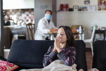 Girl sitting on a sofa blowing her nose, and a doctor seated in a face mask and white coat at a distance. - CUF55435