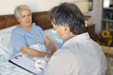 A doctor in a white coat and protective face mask making a home visit to a senior woman patient. - CUF55440