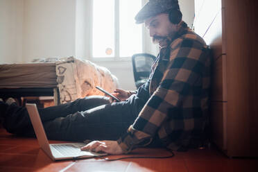 Man with big moustache wearing flat cap sitting on bedroom floor with laptop computer during Corona virus crisis. - CUF55461