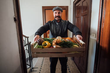 Man with big moustache wearing flat cap delivering vegetable box in an apartment building during Corona virus crisis. - CUF55469