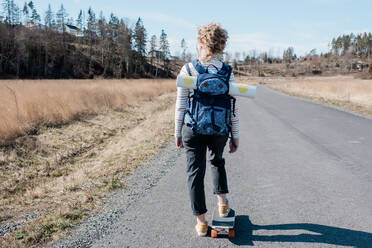 Frau trägt eine Yogamatte in einem Rucksack, während sie auf einem Skateboard skatet - CAVF81202