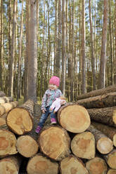 Lächelndes kleines Mädchen mit Puppe auf einem Holzstapel im Wald sitzend - BRF01457