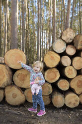 Porträt eines glücklichen kleinen Mädchens mit Puppe, das vor einem Holzstapel im Wald steht - BRF01465