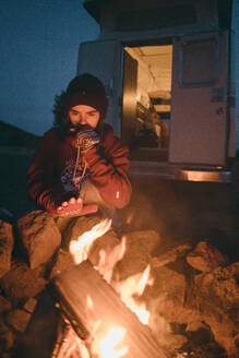 Junge Frau hält Hände in der Nähe des Feuers beim Camping in der Abenddämmerung - CAVF81307