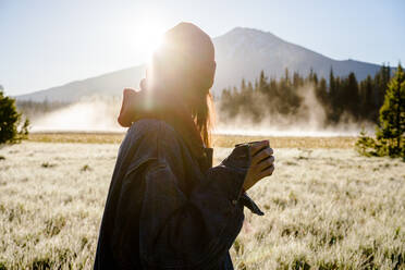 Woman at sunrise looking at foggy mountain with coffee in hand - CAVF81325