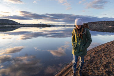 Young woman walking along lake at sunset with mountain in background - CAVF81328