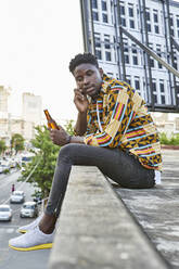 Young man in patterned shirt sitting on roof terrace in the city talking on the phone, Maputo, Mozambique - VEGF02263