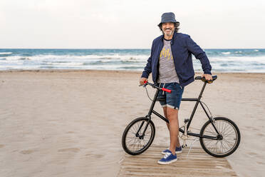 Älterer Mann mit Fahrrad, stehend am Strand, lächelnd - DLTSF00687
