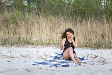 Smiling young woman sitting on the beach using smartphone and earphones - ASCF01329