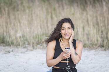 Portrait of smiling young woman on the beach using smartphone and earphones - ASCF01331
