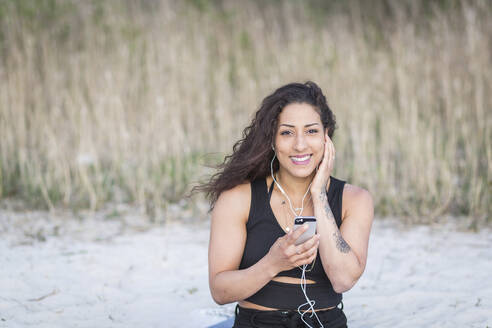 Porträt einer lächelnden jungen Frau am Strand mit Smartphone und Kopfhörern - ASCF01331