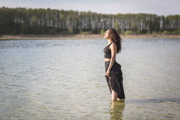 Junge Frau steht im See und genießt das Sonnenlicht, Brandenburg, Deutschland - ASCF01332