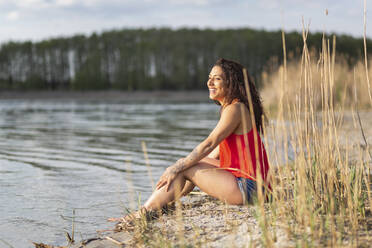 Porträt einer glücklichen jungen Frau, die am Seeufer sitzt - ASCF01336