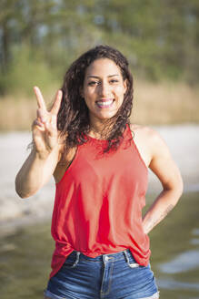Porträt einer glücklichen jungen Frau am See mit Siegeszeichen - ASCF01338
