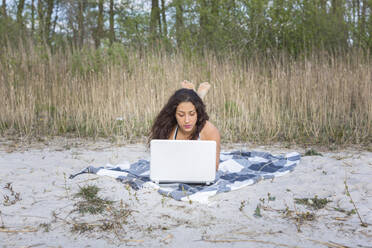 Junge Frau liegt auf einer Decke am Strand und benutzt einen Laptop - ASCF01352