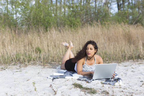 Porträt einer jungen Frau, die auf einer Decke am Strand liegt und einen Laptop benutzt - ASCF01354