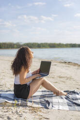 Young woman sitting on blanket on the beach using laptop - ASCF01356