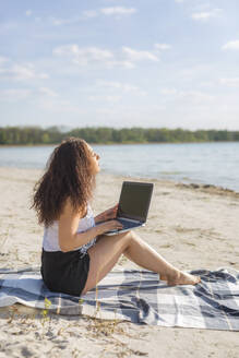 Junge Frau sitzt auf einer Decke am Strand und benutzt einen Laptop - ASCF01356