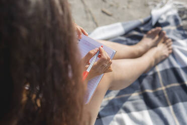 Ansicht einer jungen Frau, die auf einer Decke am Strand sitzt und in ein Notizbuch schreibt - ASCF01358