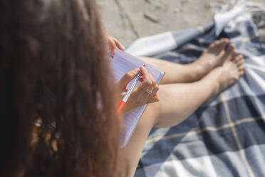 Crop view of young woman sitting on blanket on the beach writing in notebook - ASCF01358