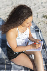 Junge Frau sitzt auf einer Decke am Strand und schreibt in ein Notizbuch - ASCF01359