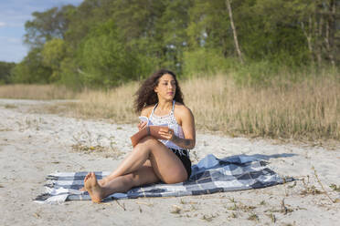 Junge Frau mit Notebook sitzt auf einer Decke am Strand und schaut in die Ferne - ASCF01360