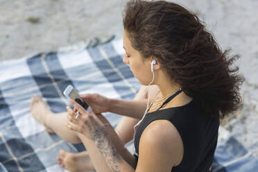 Young woman sitting on blanket on the beach using earphones and smartphone - ASCF01362