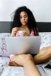 Schöne junge Frau im Bett liegend zu Hause mit einer Tasse Kaffee und Laptop - MGOF04315