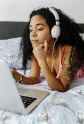 Schöne junge Frau liegt zu Hause im Bett, trägt Kopfhörer und benutzt einen Laptop - MGOF04320
