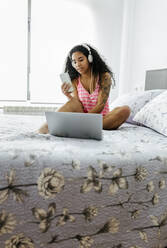 Schöne junge Frau mit Kopfhörern, Smartphone und Laptop im Bett - MGOF04322