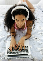 Schöne junge Frau liegt zu Hause im Bett, trägt Kopfhörer und benutzt einen Laptop - MGOF04323
