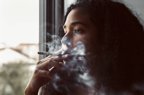 Porträt einer jungen Frau, die am Fenster eine Zigarette raucht - MGOF04334