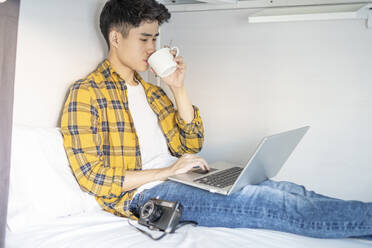 Junger Tourist sitzt auf einem Bett in einer Herberge und trinkt eine Tasse Kaffee, während er einen Laptop benutzt - JCZF00072