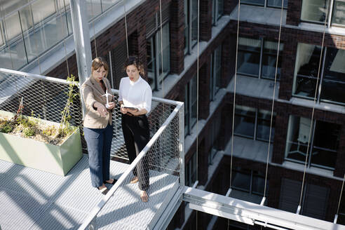 Two businesswomen with tablet and wind turbine model having a meeting on balcony of an office building - JOSEF00661