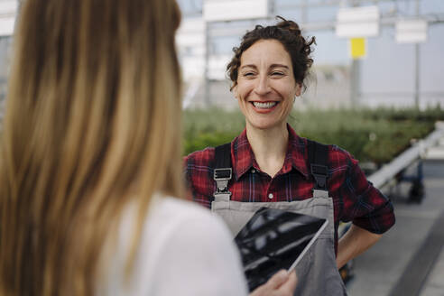 Smiling gardener looking at businesswoman in greenhouse of a gardening shop - JOSEF00671