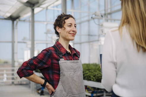 Smiling gardener looking at businesswoman in greenhouse of a gardening shop - JOSEF00672