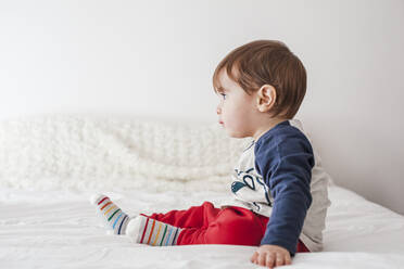 Seitenansicht eines kleinen Jungen, der auf dem Bett sitzt und in die Ferne schaut - FLMF00219