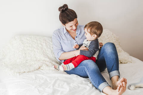 Glückliche Mutter sitzt auf dem Bett und spielt mit ihrem kleinen Sohn, lizenzfreies Stockfoto