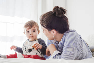 Porträt eines kleinen Jungen, der auf dem Bett sitzt und Kekse isst, während seine Mutter ihn beobachtet - FLMF00224