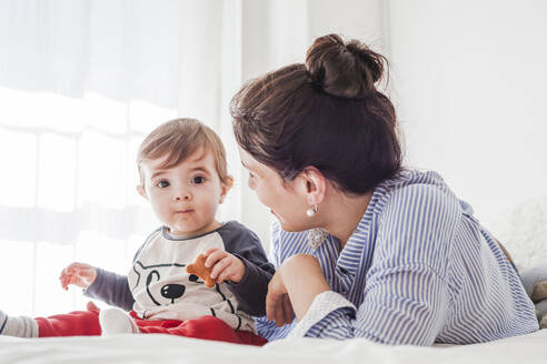 Portrait of baby boy sitting on bed eating cookie while his mother watching him - FLMF00224