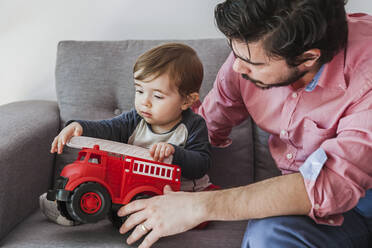 Kleiner Junge sitzt mit seinem Vater auf der Couch und spielt mit einem Feuerwehrauto - FLMF00226