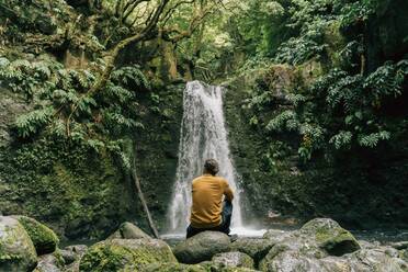 Rückansicht eines Mannes, der an einem Wasserfall auf der Insel Sao Miguel, Azoren, Portugal, sitzt - AFVF06289